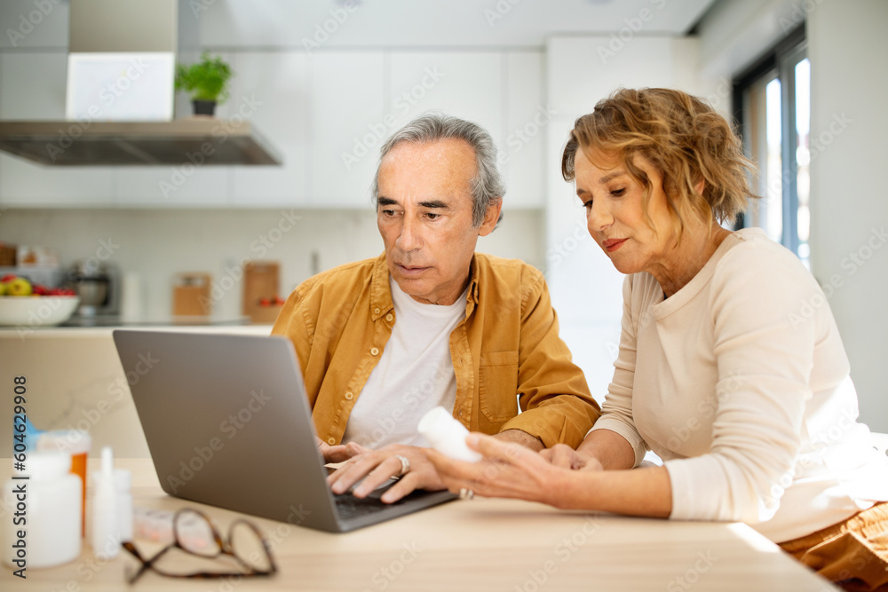 Senior couple holding jar of pills and searching prescription with help of laptop computer, sitting at table in kitchen Senior couple holding jar of pills and searching prescription with help of laptop computer, sitting at table in kitchen