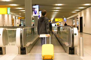  Young teen boy with yellow suitcase from back walking along the walk way in international airport to the gate. Teenager travel alone concept.