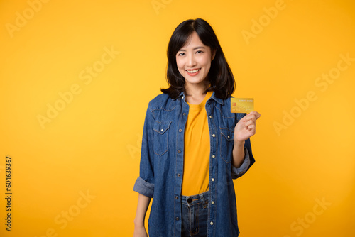 Portrait of young asian woman wearing yellow t-shirt and denim jacket with happy smile holding a credit card isolated on yellow background. Payment shopping online concept.