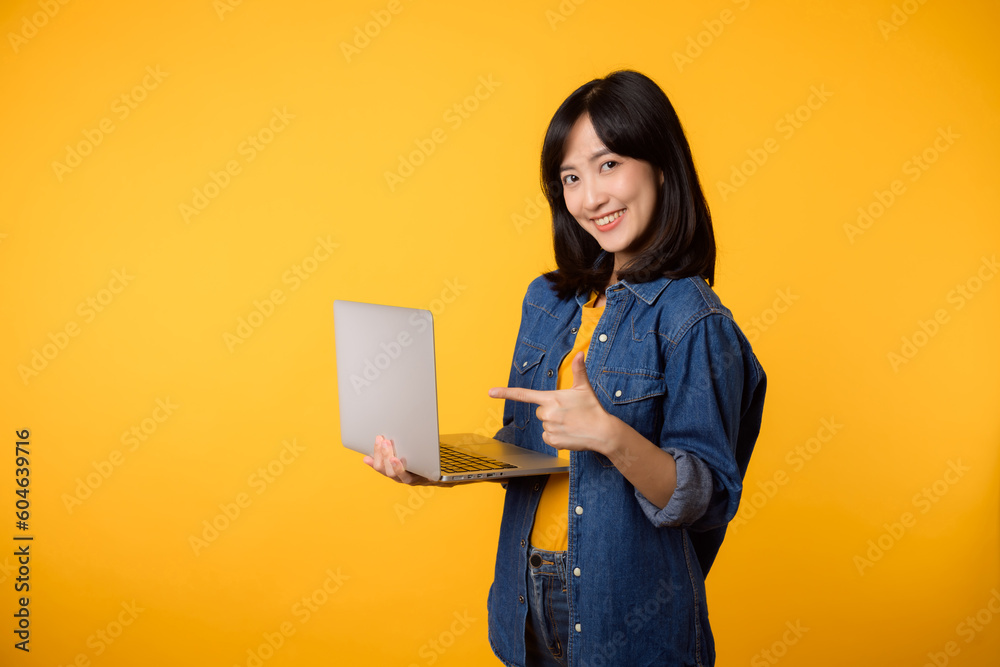 portrait young happy woman wearing yellow t-shirt and denim shirt holding laptop and point finger to screen isolated on yellow studio background. business technology application communication concept. portrait young happy woman wearing yellow t-shirt and denim shirt holding laptop and point finger to screen isolated on yellow studio background. business technology application communication concept.