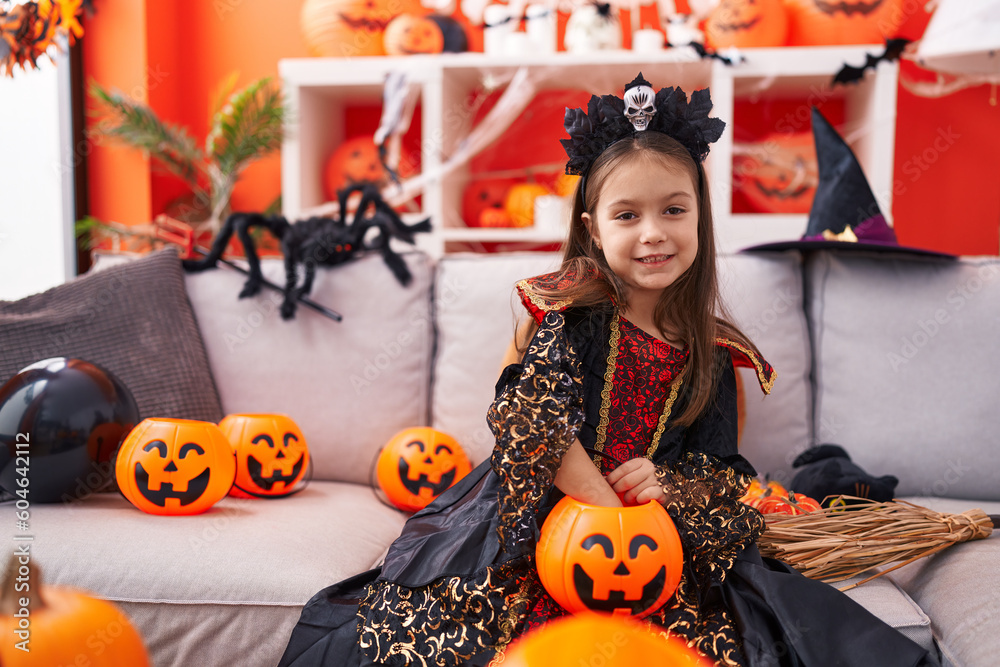 Fototapeta premium Adorable hispanic girl wearing halloween costume holding candy of pumpkin basket at home