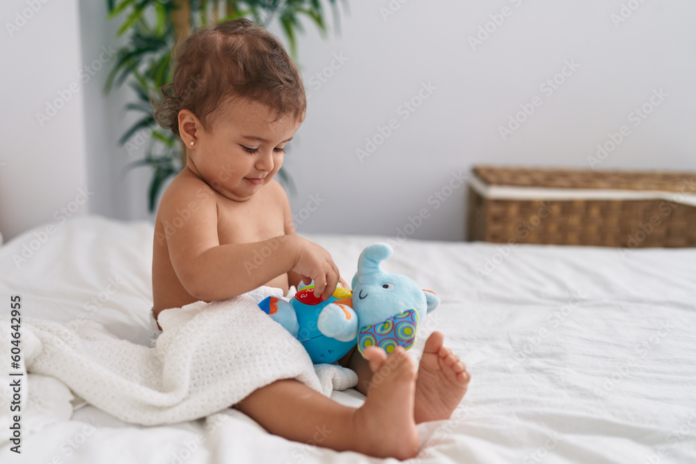 Adorable hispanic baby sitting on bed playing with elephant doll at bedroom