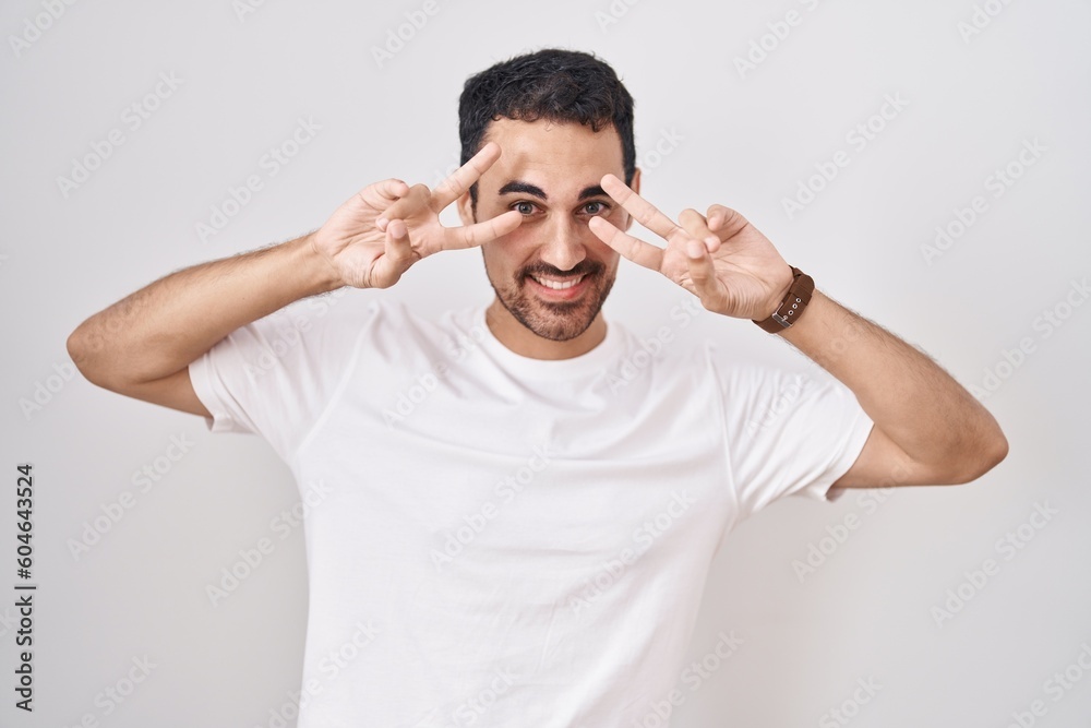 Handsome hispanic man standing over white background doing peace symbol with fingers over face, smiling cheerful showing victory Handsome hispanic man standing over white background doing peace symbol with fingers over face, smiling cheerful showing victory
