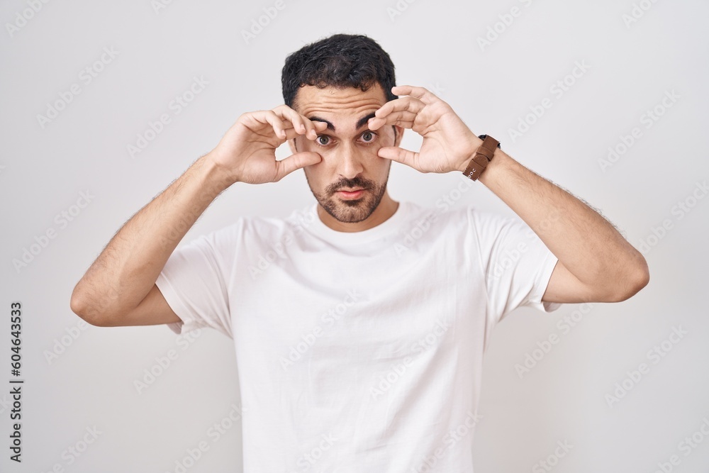 Handsome hispanic man standing over white background trying to open eyes with fingers, sleepy and tired for morning fatigue Handsome hispanic man standing over white background trying to open eyes with fingers, sleepy and tired for morning fatigue