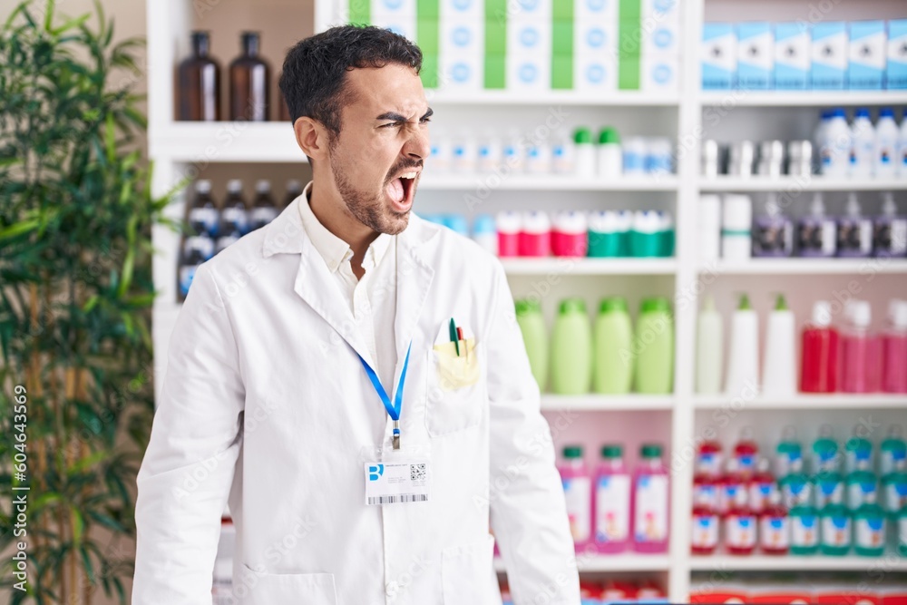 Handsome hispanic man working at pharmacy drugstore angry and mad screaming frustrated and furious, shouting with anger. rage and aggressive concept. Handsome hispanic man working at pharmacy drugstore angry and mad screaming frustrated and furious, shouting with anger. rage and aggressive concept.