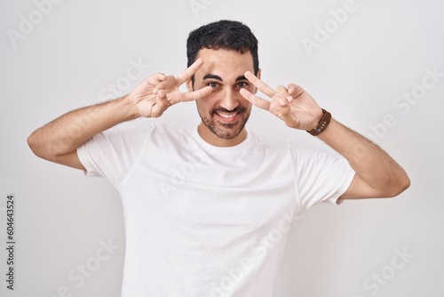 Handsome hispanic man standing over white background doing peace symbol with fingers over face, smiling cheerful showing victory © Krakenimages.com