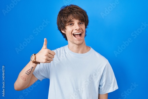 Hispanic young man standing over blue background doing happy thumbs up gesture with hand. approving expression looking at the camera showing success.