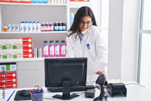 Young hispanic girl pharmacist talking by telephone reading document at pharmacy