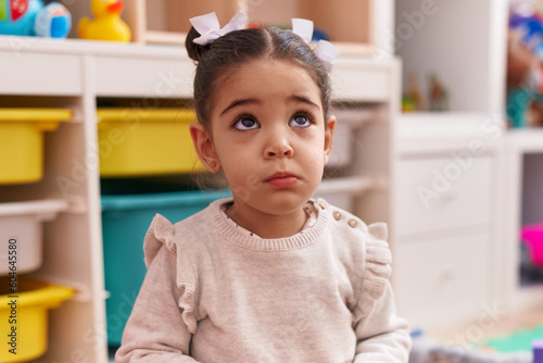 Adorable hispanic girl sitting on floor with sad expression at kindergarten