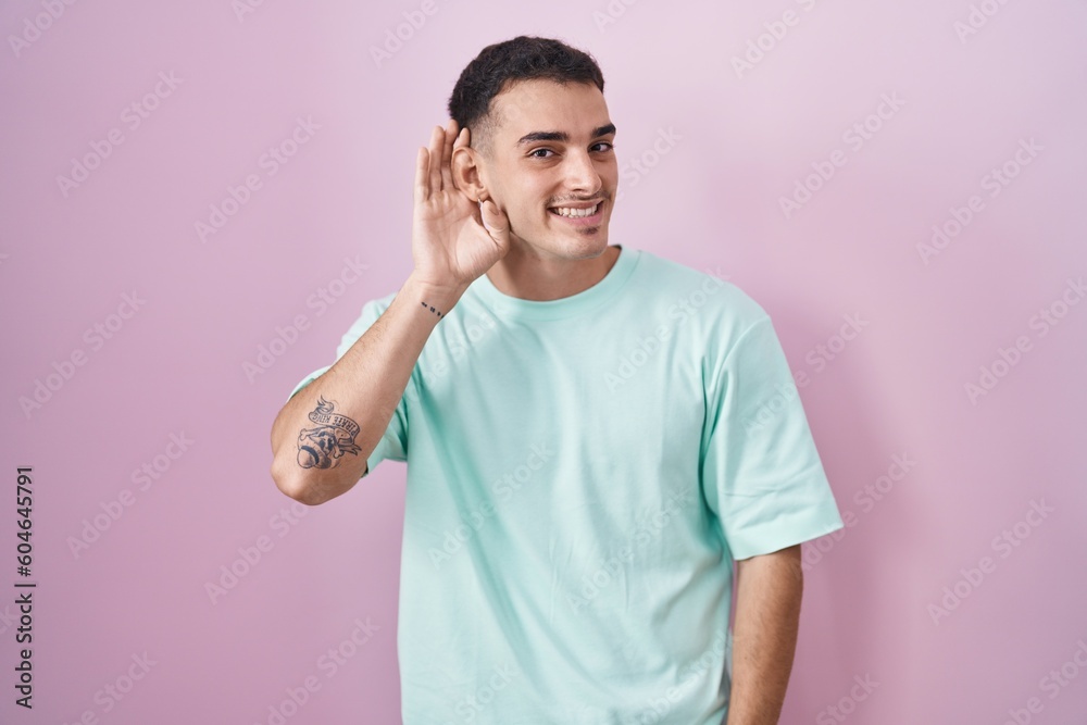 Handsome hispanic man standing over pink background smiling with hand over ear listening an hearing to rumor or gossip. deafness concept. Handsome hispanic man standing over pink background smiling with hand over ear listening an hearing to rumor or gossip. deafness concept.