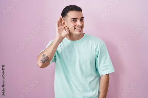 Handsome hispanic man standing over pink background smiling with hand over ear listening an hearing to rumor or gossip. deafness concept.