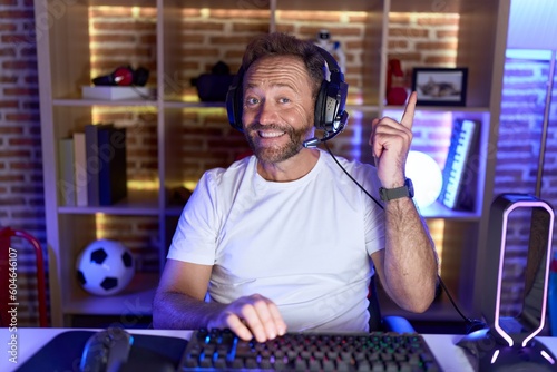 Middle age man with beard playing video games wearing headphones with a big smile on face, pointing with hand finger to the side looking at the camera.