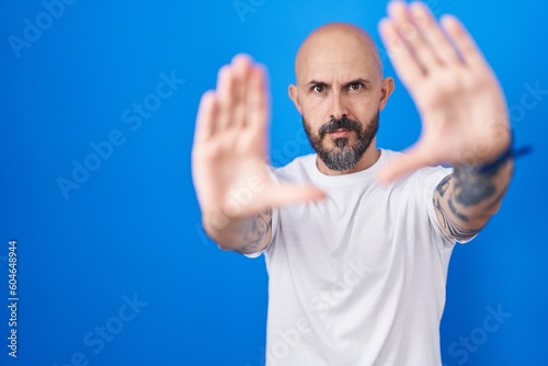 Hispanic man with tattoos standing over blue background doing frame using hands palms and fingers, camera perspective