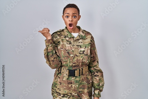 Beautiful african american woman wearing camouflage army uniform surprised pointing with hand finger to the side  open mouth amazed expression.
