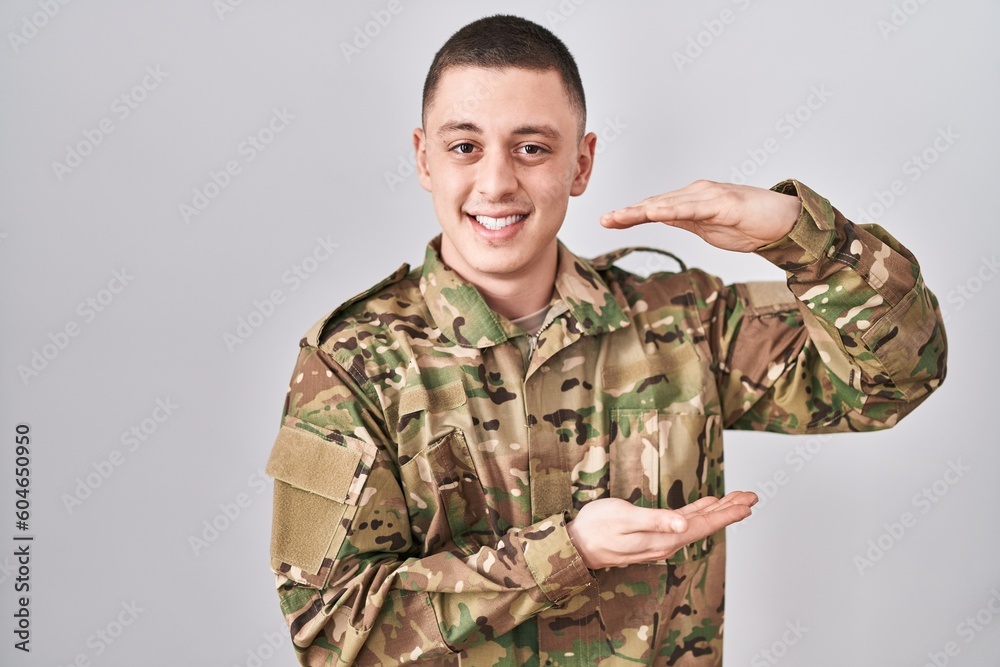 Young man wearing camouflage army uniform gesturing with hands showing big and large size sign, measure symbol. smiling looking at the camera. measuring concept.