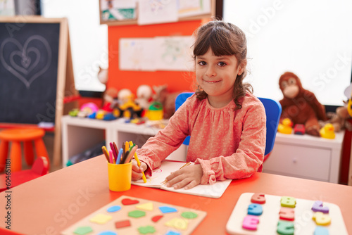 Adorable hispanic girl student sitting on table drawing on notebook at kindergarten