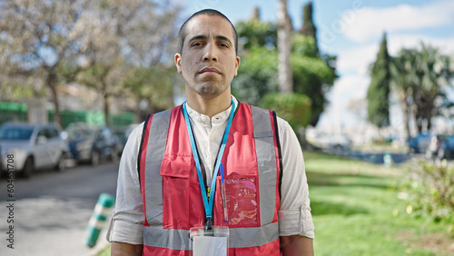 Young hispanic man volunteer wearing vest looking serious at park