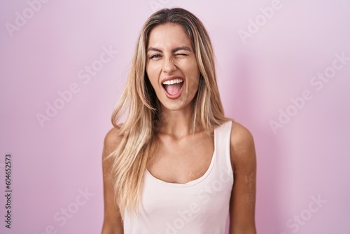 Young blonde woman standing over pink background winking looking at the camera with sexy expression, cheerful and happy face.