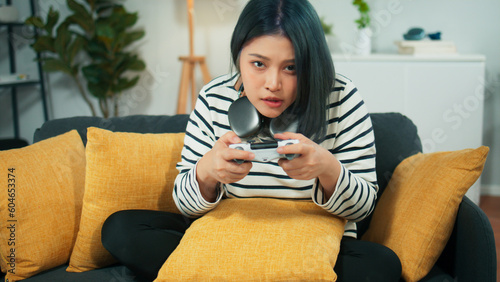 Excited beautiful Asian woman using joystick wireless controller enjoy playing fighting console video game to win highest victory. Girl Gamer sit on sofa in modern living room at home
