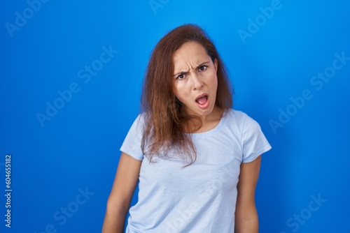 Brunette woman standing over blue background in shock face, looking skeptical and sarcastic, surprised with open mouth