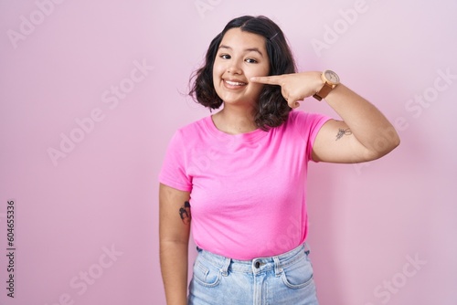 Young hispanic woman standing over pink background pointing with hand finger to face and nose, smiling cheerful. beauty concept