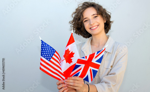 Smiling student woman hold Canada, UK and US flags and looking camera on gray wall background. English language education.
