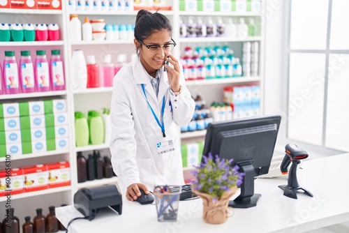 Young beautiful hispanic woman pharmacist talking on smartphone using computer at pharmacy