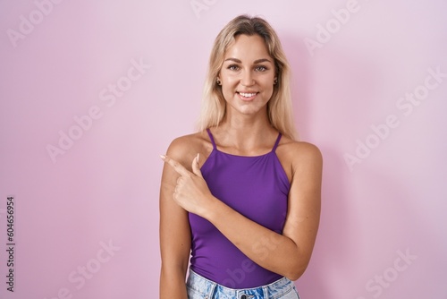 Young blonde woman standing over pink background cheerful with a smile on face pointing with hand and finger up to the side with happy and natural expression