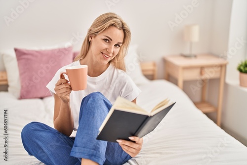 Young blonde woman drinking cup of coffee reading book at bedroom
