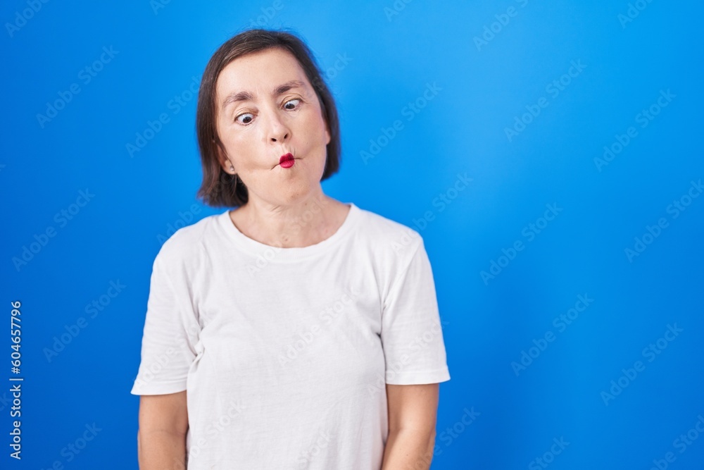 Middle age hispanic woman standing over blue background making fish face with lips, crazy and comical gesture. funny expression. Middle age hispanic woman standing over blue background making fish face with lips, crazy and comical gesture. funny expression.