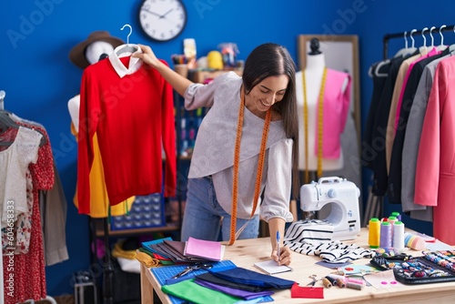 Young beautiful hispanic woman tailor smiling confident holding t shirt writing on notebook at clothing factory
