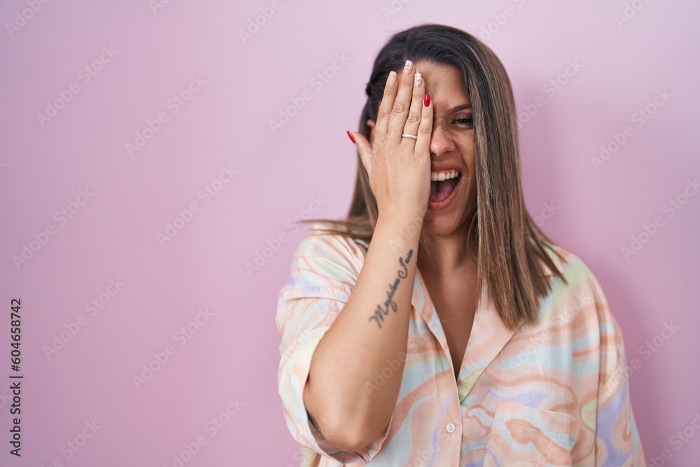 Blonde woman standing over pink background covering one eye with hand, confident smile on face and surprise emotion. Blonde woman standing over pink background covering one eye with hand, confident smile on face and surprise emotion.