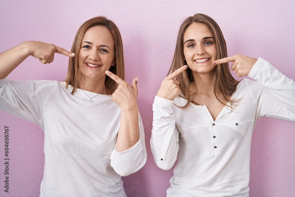 Middle age mother and young daughter standing over pink background smiling cheerful showing and pointing with fingers teeth and mouth. dental health concept. Middle age mother and young daughter standing over pink background smiling cheerful showing and pointing with fingers teeth and mouth. dental health concept.