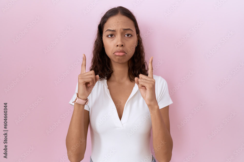 Young hispanic woman wearing casual white t shirt pointing up looking sad and upset, indicating direction with fingers, unhappy and depressed. Young hispanic woman wearing casual white t shirt pointing up looking sad and upset, indicating direction with fingers, unhappy and depressed.
