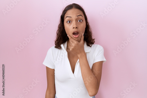 Young hispanic woman wearing casual white t shirt looking fascinated with disbelief, surprise and amazed expression with hands on chin