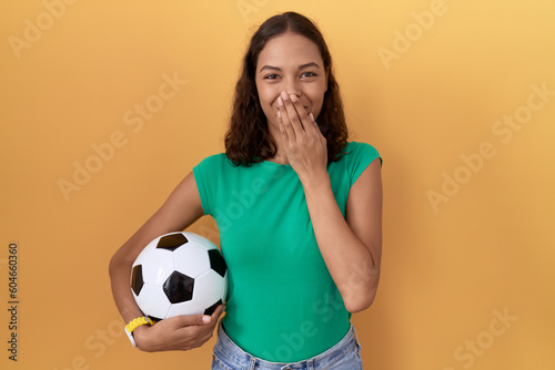 Young hispanic woman holding ball laughing and embarrassed giggle covering mouth with hands, gossip and scandal concept © Krakenimages.com