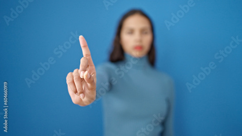 Young beautiful hispanic woman saying no with finger over isolated blue background photo