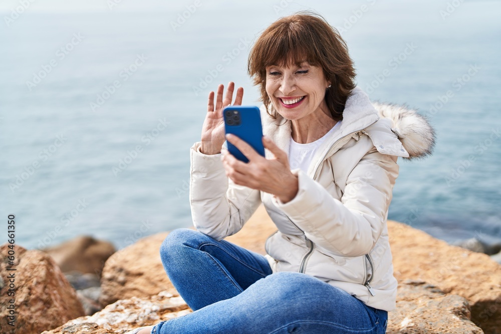 Middle age woman having video call sitting on the rock at seaside