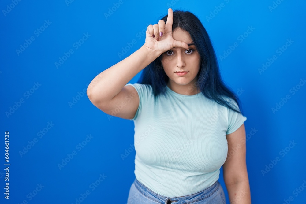 Young modern girl with blue hair standing over blue background making fun of people with fingers on forehead doing loser gesture mocking and insulting.