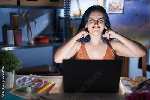 Young modern girl with blue hair sitting at art studio with laptop at night covering ears with fingers with annoyed expression for the noise of loud music. deaf concept.