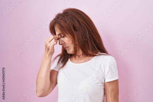 Middle age woman standing over pink background tired rubbing nose and eyes feeling fatigue and headache. stress and frustration concept.