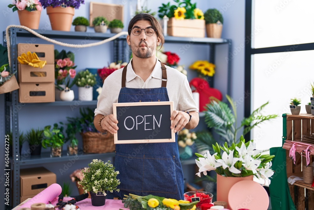 Hispanic man with long hair working at florist holding open sign making fish face with mouth and squinting eyes, crazy and comical.