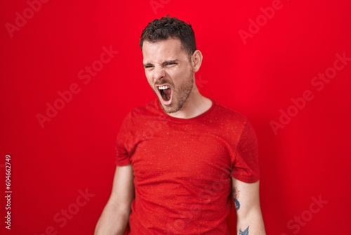 Young hispanic man standing over red background angry and mad screaming frustrated and furious, shouting with anger. rage and aggressive concept. © Krakenimages.com