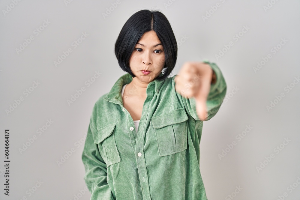 Young asian woman standing over white background looking unhappy and angry showing rejection and negative with thumbs down gesture. bad expression. Young asian woman standing over white background looking unhappy and angry showing rejection and negative with thumbs down gesture. bad expression.
