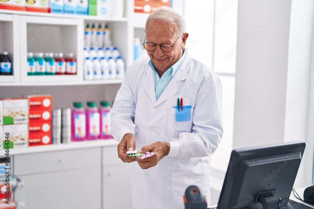 Senior grey-haired man pharmacist smiling confident holding pills tablets at pharmacy