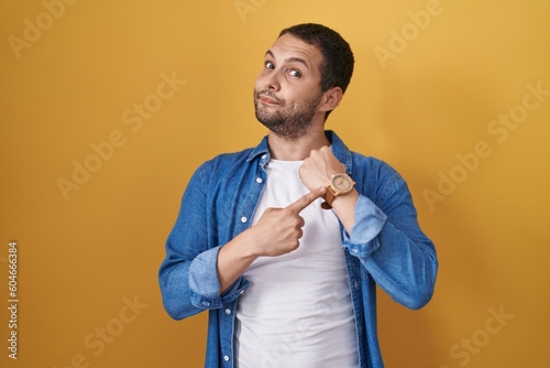 Hispanic man standing over yellow background in hurry pointing to watch time, impatience, looking at the camera with relaxed expression photo