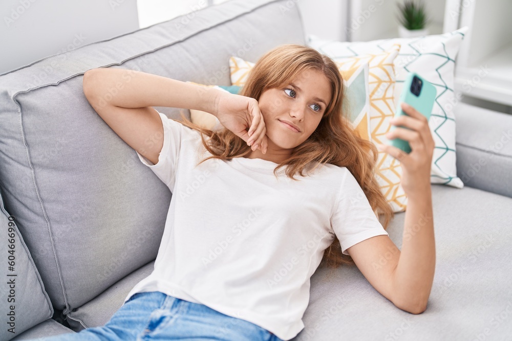 Young blonde girl using smartphone lying on sofa at home