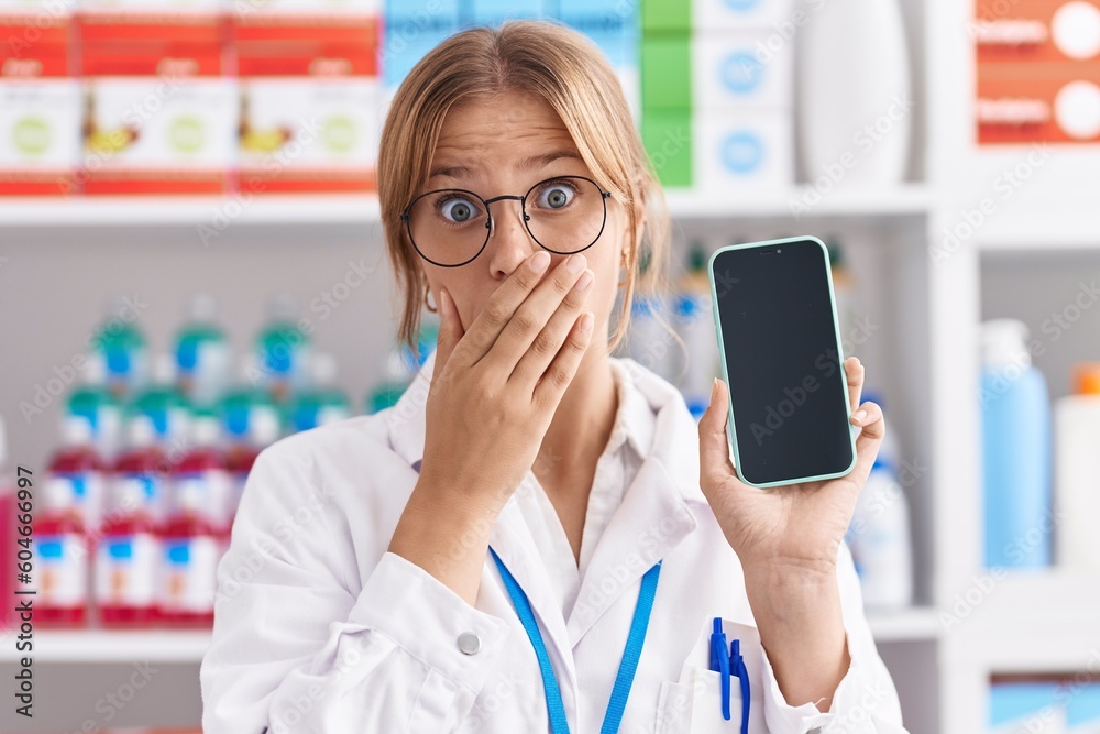 Young caucasian woman working at pharmacy drugstore showing smartphone screen covering mouth with hand, shocked and afraid for mistake. surprised expression Young caucasian woman working at pharmacy drugstore showing smartphone screen covering mouth with hand, shocked and afraid for mistake. surprised expression