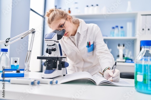 Young blonde woman scientist using microscope writing report at laboratory