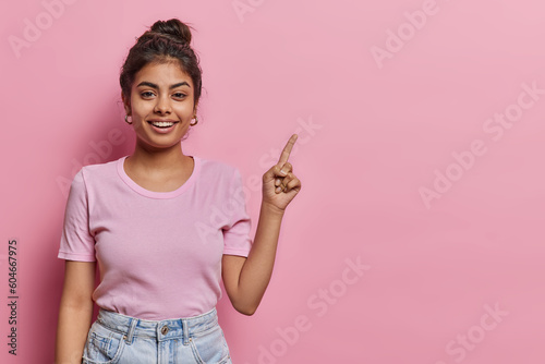 Attractive Indian teenage girl with dark hair points index finger above on copy space shows promotion offer demonstrates advertisement dressed in casual t shirt and jeans isolated over pink background